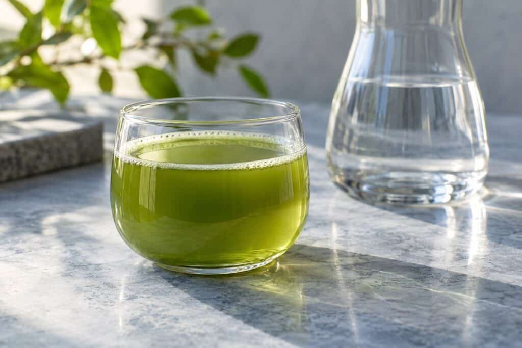 كوب شفاف من شاي الماتشا الأخضر بجانب دورق ماء على طاولة حجرية شاحبة