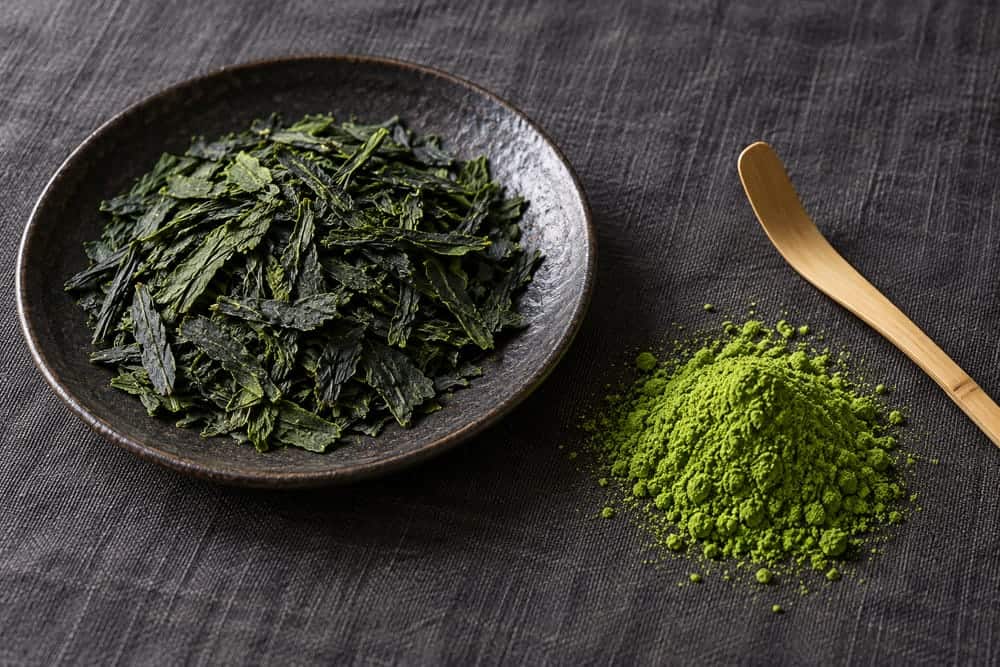 Dried tea leaf flakes beside fine matcha powder and a bamboo scoop